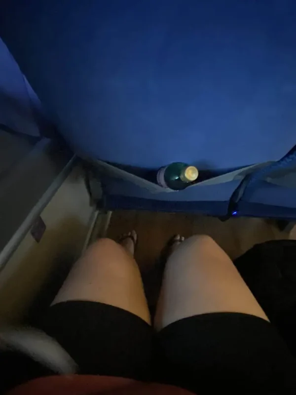 A passenger's view inside a bus traveling from Cancun to Caye Caulker. The image shows a pair of knees in shorts, a bottle of water in the seat pocket, and a headrest with a paper cover labeled 'ADO'. The bus interior includes blue seats and ample legroom.