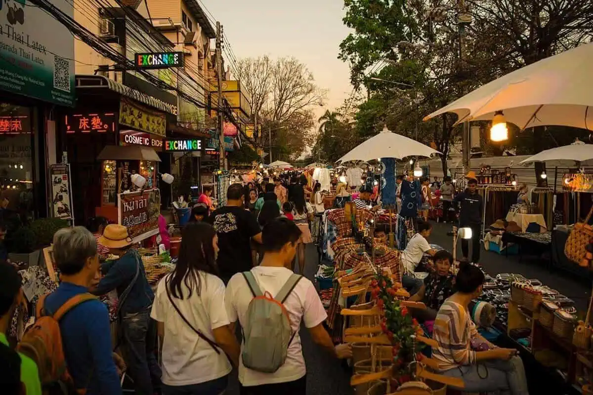 Shoppers and vendors on a busy street with stalls offering a variety of goods during golden hour.