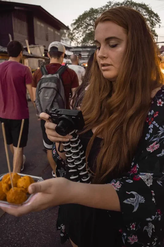 Tasha Amy at the Sunday night market in Chiang Mai focused on her camera while holding a plate of Thai fried balls, with market-goers in the background.