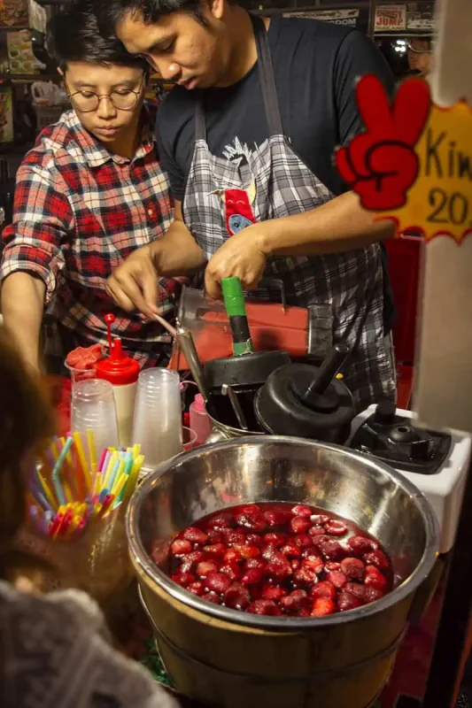 Vendors preparing drinks at a stall with a colorful display of straws and a large bowl of strawberries.