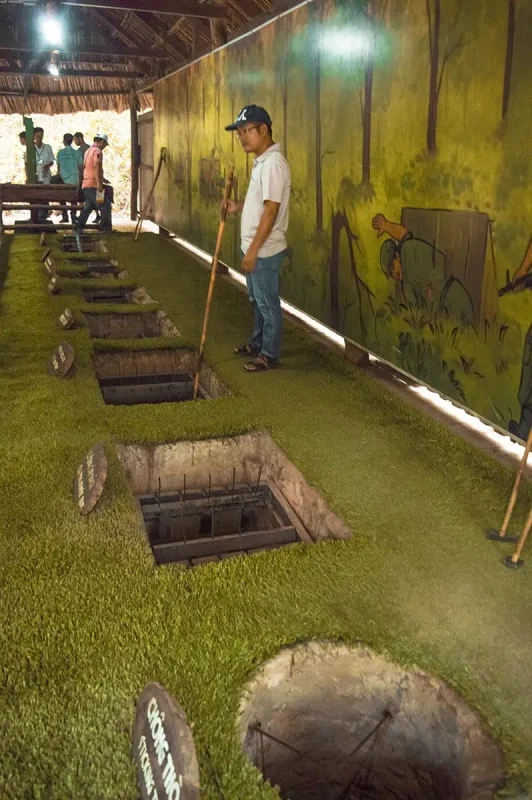 Best Cu Chi Tunnels Tours: Cheap VS Expensive 2025 11 An interior view of a Cu Chi Tunnels exhibit with various open trapdoors in the ground, demonstrating guerrilla tactics with a guide and visitors in the background, painted walls depict a jungle environment.