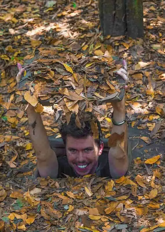 Best Cu Chi Tunnels Tours: Cheap VS Expensive 2025 7 A man smiling joyfully while emerging from a Cu Chi Tunnel camouflaged with a cover of dry leaves, with the dense forest around him.
