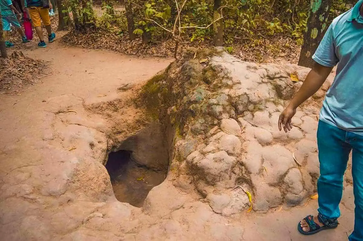 Best Cu Chi Tunnels Tours: Cheap VS Expensive 2025 9 A man stands beside a small, open tunnel entrance at the Cu Chi Tunnels, with visitors in the background walking through the forested area.