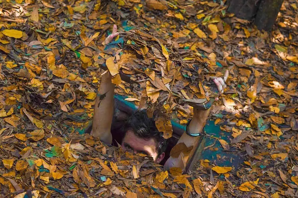 Best Cu Chi Tunnels Tours: Cheap VS Expensive 2025 10 A man peers out of a Cu Chi tunnel, concealed by a camouflage of dry leaves, illustrating the stealthy design of the tunnels.