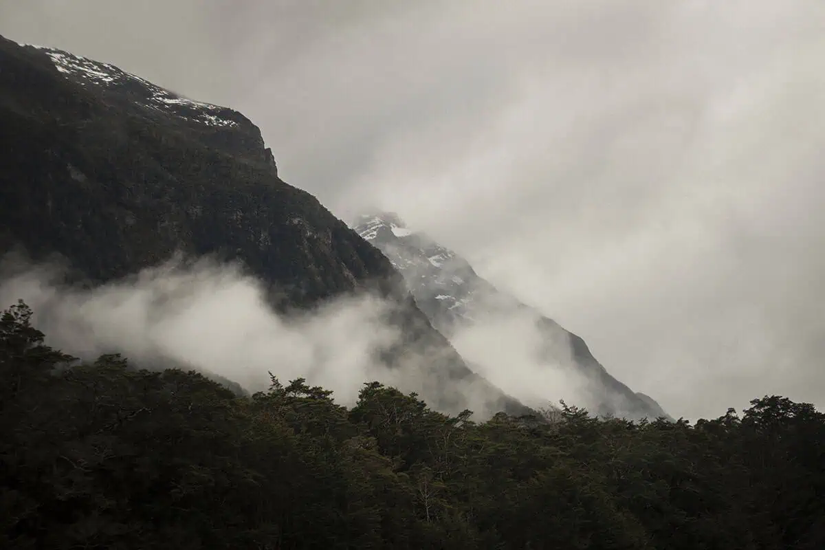 Close-up view of mist and clouds clinging to the steep, rugged slopes of a mountain partially covered in snow, creating a dramatic and mysterious atmosphere.