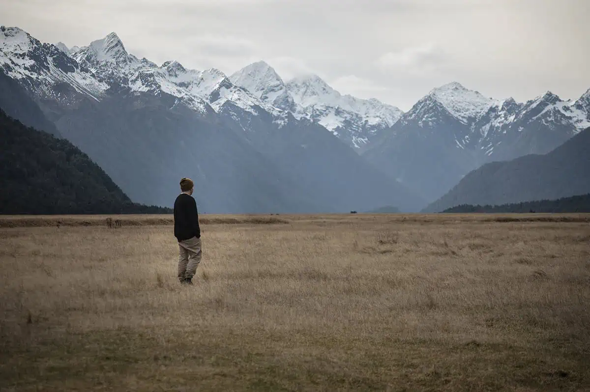 A man gazes into the distance while standing in a wide expanse of dry grass, with towering snow-capped mountains creating a breathtaking backdrop. The tranquil setting of Eglinton Valley emphasizes the natural beauty of the area.