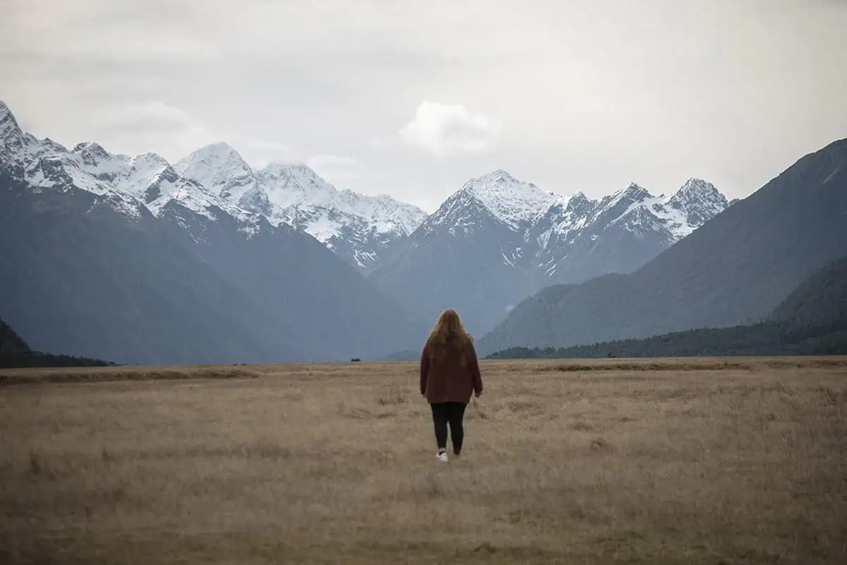 tasha amy with long hair walks away from the camera across an expansive field, with the dramatic, snow-capped peaks of Eglinton Valley rising in the distance under a cloudy sky.