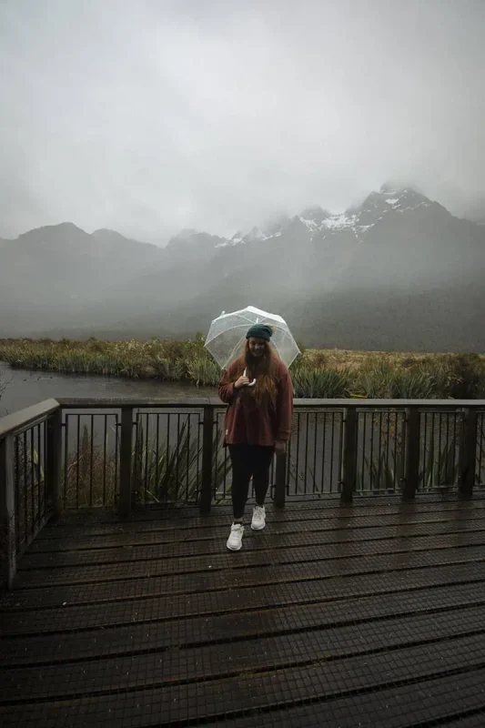 tasha amy stands on a wooden platform holding a clear umbrella, with misty mountains and a river in the background. The overcast weather adds a mystical charm to the scenic landscape of Eglinton Valley.