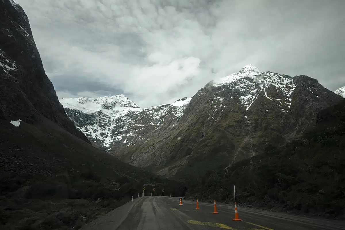 A winding road flanked by steep, rugged mountains with patches of snow, leading into the distance under a cloudy sky. The dramatic landscape driving to Milford Sound is highlighted by the stark contrast between the road and the surrounding wilderness