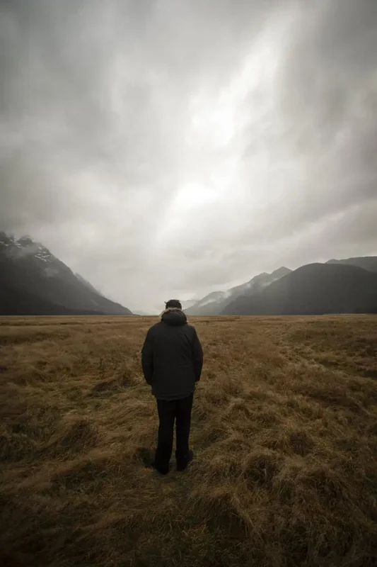 A person in a dark coat stands facing away, looking towards the expansive plains and misty mountains in the distance. The overcast sky and muted tones create a reflective and tranquil mood in Eglinton Valley.