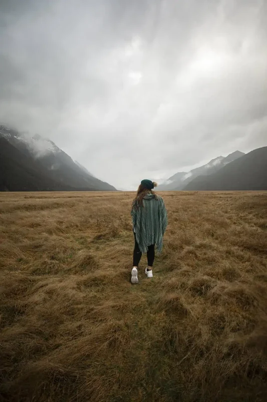 tasha amy dressed in a green poncho and white sneakers walks across the dry grass, surrounded by towering mountains and an overcast sky. The misty peaks in the distance add to the dramatic scenery of Eglinton Valley.