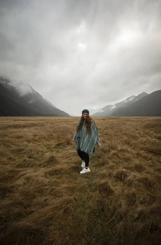 tasha amy in a green poncho smiles while standing on a vast, grassy plain, with mist-covered mountains in the background. The scene captures the serene and breathtaking landscape of Eglinton Valley.