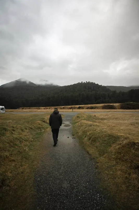 A person in a dark coat walks along a gravel path leading into the distance, bordered by grassy fields and distant forested hills under a cloudy sky. The peaceful solitude of the scene is accentuated by the misty weather.
