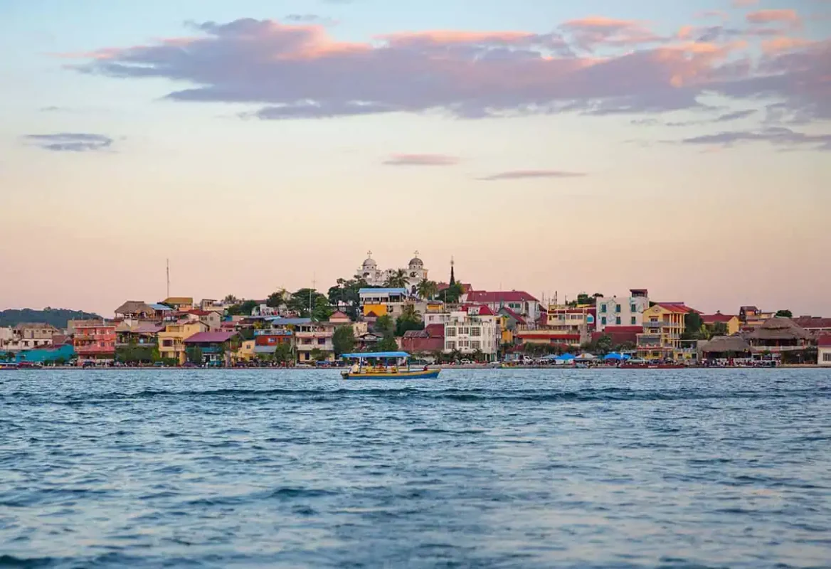The vibrant town of Flores is seen from a distance, with colorful buildings lining the waterfront against a pastel sunset sky. A boat travels across the calm waters, capturing the serene atmosphere of this picturesque Guatemalan town. This image represents the starting point for a journey from Flores to Belize City.