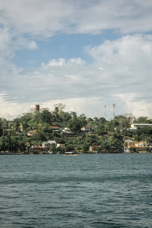 The shore of a body of water in Flores, with lush greenery and houses on the hillside under a partly cloudy sky. This serene waterfront scene highlights the natural beauty of Flores.