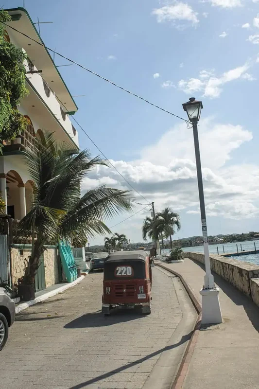 A coastal road in Flores, featuring a small red vehicle with the number 220 parked beside a lamppost and palm trees. The image captures the relaxed coastal vibe, typical of Flores.