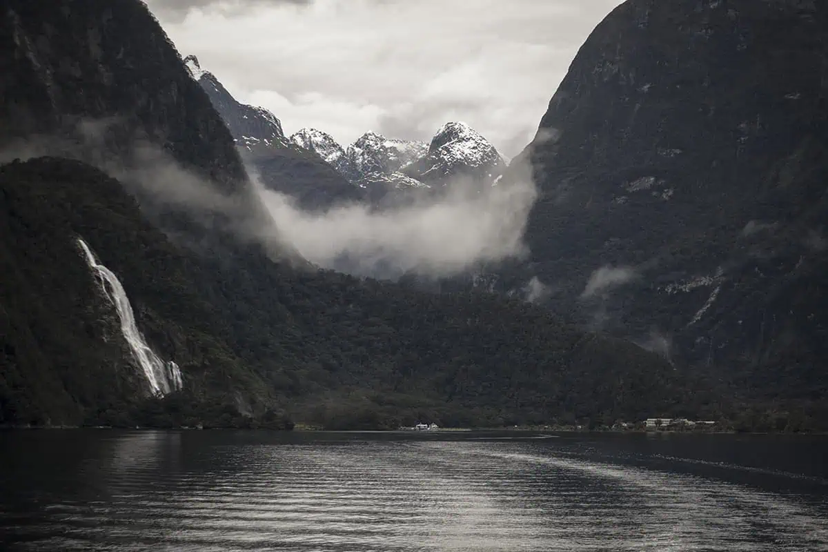 Snowy mountains and a waterfall framed by misty cliffs when looking back towards the small township in Milford Sound.