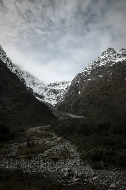 looking up a dried up stream towards the snowy mountains when driving into milford sound