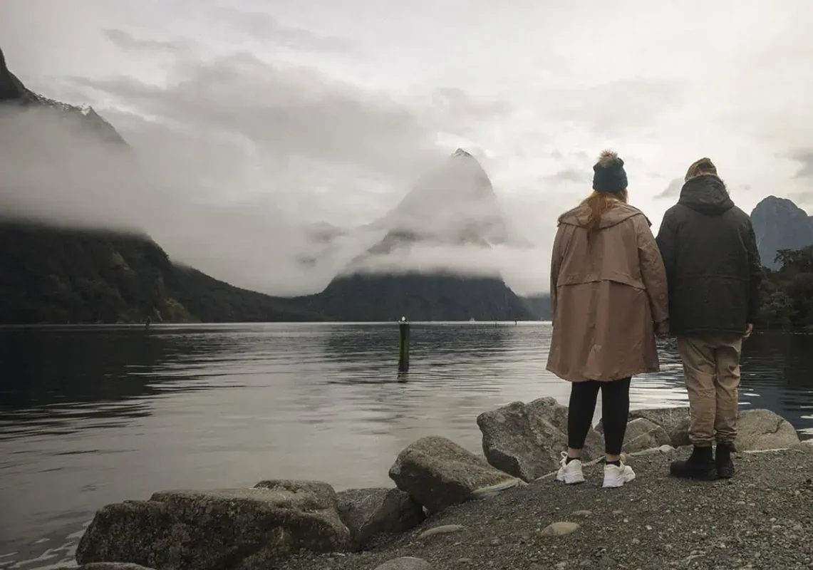 A couple standing hand-in-hand on the shoreline, gazing over the water to Mitre Peak.