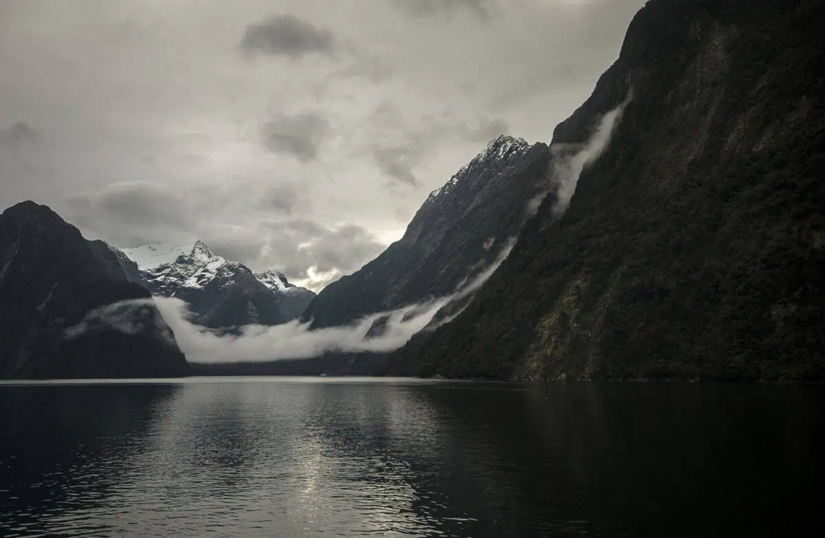 Snow covered peaks and low clouds reflected in the calm waters of Milford Sound.