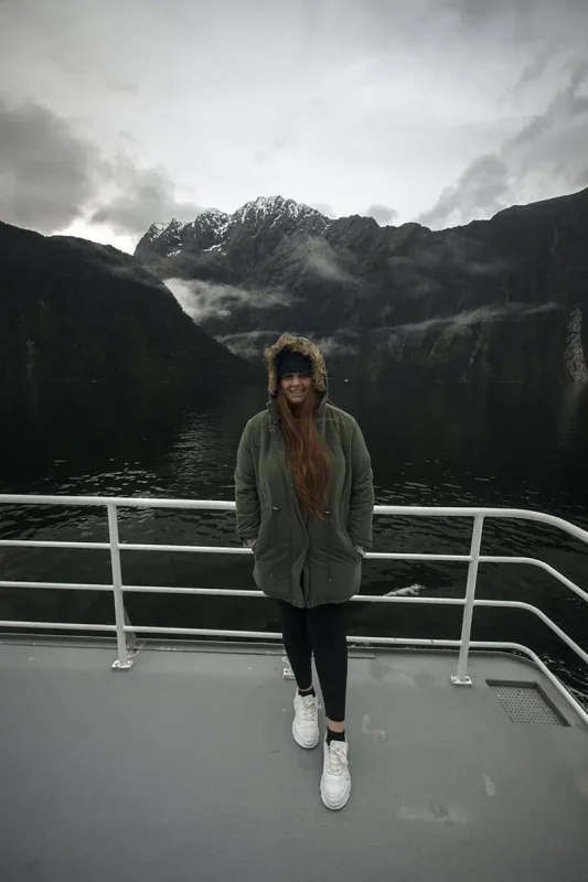 tasha amy in a winter coat on the deck of a Milford Sound cruise with mountains in the background.