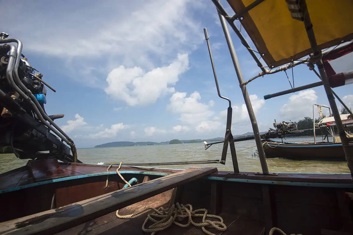View from a long-tail boat on Railay Beach, showing the engine, a coiled rope, and the expansive sea under a bright blue sky with scattered clouds. This image suggests boating activities are ongoing, posing the question: "Is Railay Beach closed?"