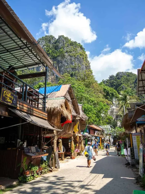 Lively street scene at Railay Beach with tourists walking past shops and cafes, set against the backdrop of lush green hills and a clear blue sky.