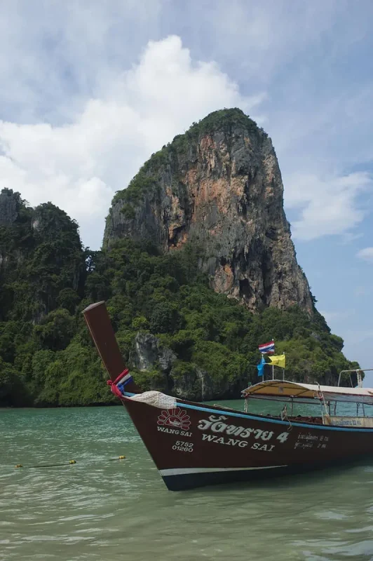 A long-tail boat named "Wang Sai" anchored near a towering limestone cliff covered in greenery at Railay Beach. The presence of boats and tourists hints at the beach being operational