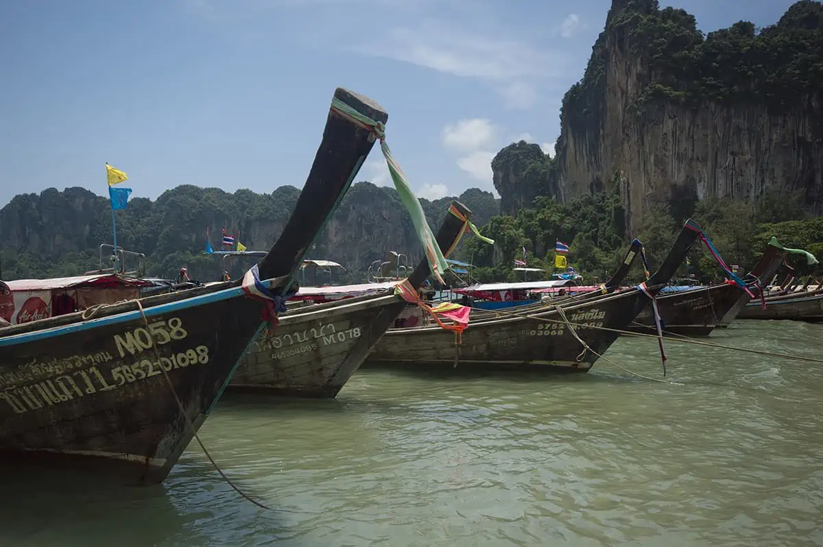 Several long-tail boats with colorful flags are lined up in the water with towering cliffs in the background at Railay Beach. This busy scene implies active tourist engagement, questioning if Railay Beach is closed.