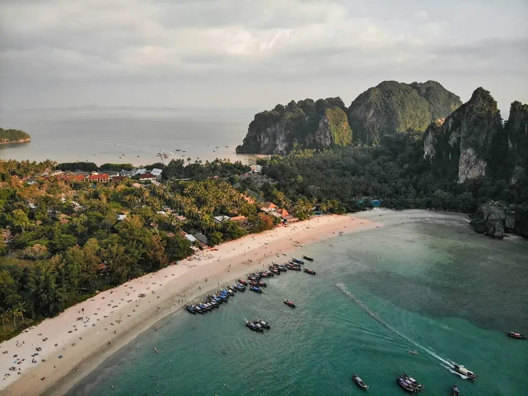 Aerial view of Railay Beach with sandy shores, numerous boats docked near the water, and lush greenery surrounding the area under a bright sky.
