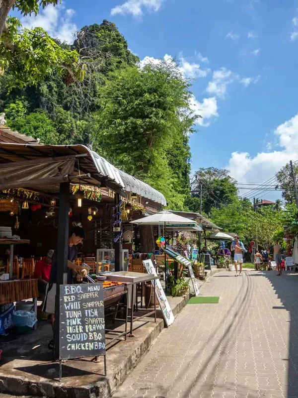 A sunny street on Railay Beach lined with small shops and restaurants, featuring a chalkboard menu offering seafood BBQ options like red snapper and tiger prawn.