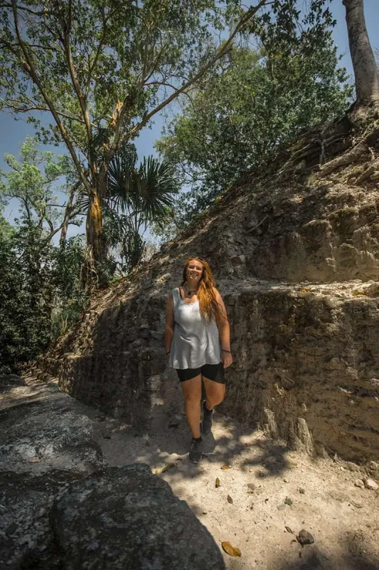 Mexico And Belize Itinerary: From 2 to 5 Weeks 49 tasha amystanding and smiling on a shaded path beside ancient Mayan ruins surrounded by dense tropical forest. She is wearing a sleeveless top and shorts, capturing the essence of adventure and exploration in belize.