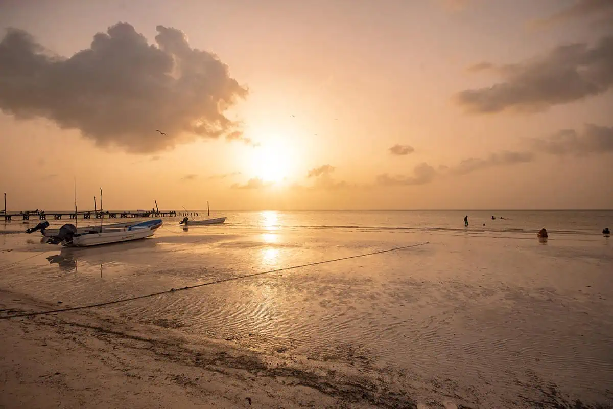 Mexico And Belize Itinerary: From 2 to 5 Weeks 7 Sunset on an isla holbox beach with small boats anchored in shallow water. The sky is painted with warm hues of orange and pink, reflecting on the water, creating a serene and picturesque moment.
