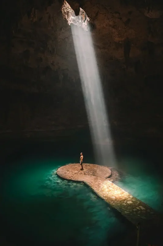 Mexico And Belize Itinerary: From 2 to 5 Weeks 23 A person standing on a stone platform inside a dark cenote, illuminated by a dramatic beam of sunlight from an opening above. The clear turquoise water surrounding the platform adds to the mystical and awe-inspiring atmosphere of this natural wonder.