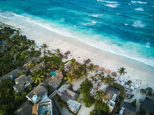 Aerial view of a picturesque beach in Mexico with clear turquoise waters and gentle waves meeting a pristine sandy shore, the perfect place to visit on a mexico and belize itinerary. The beach is lined with palm trees and several thatched-roof huts, some with small swimming pools, offering a tranquil and inviting tropical getaway.
