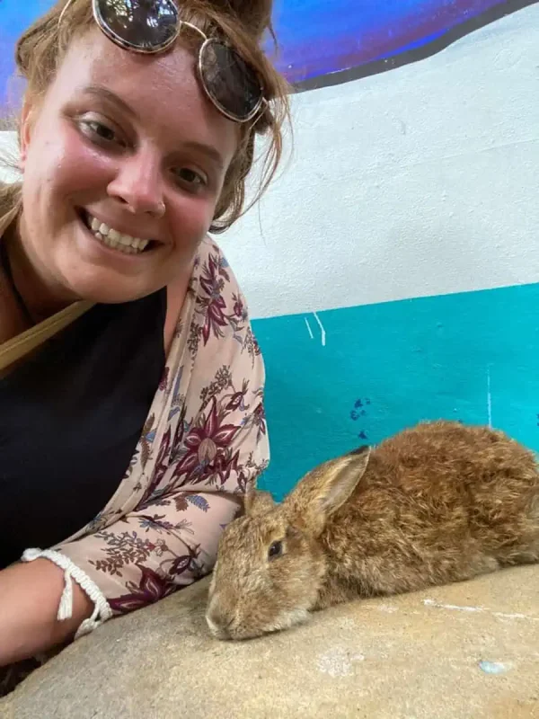 A close-up image of tasha amy smiling at the camera, with a brown rabbit resting beside her against a teal and white wall. She wears a floral-patterned cover-up and sunglasses atop her head, sharing a moment of joy with the rabbit in a casual setting.
