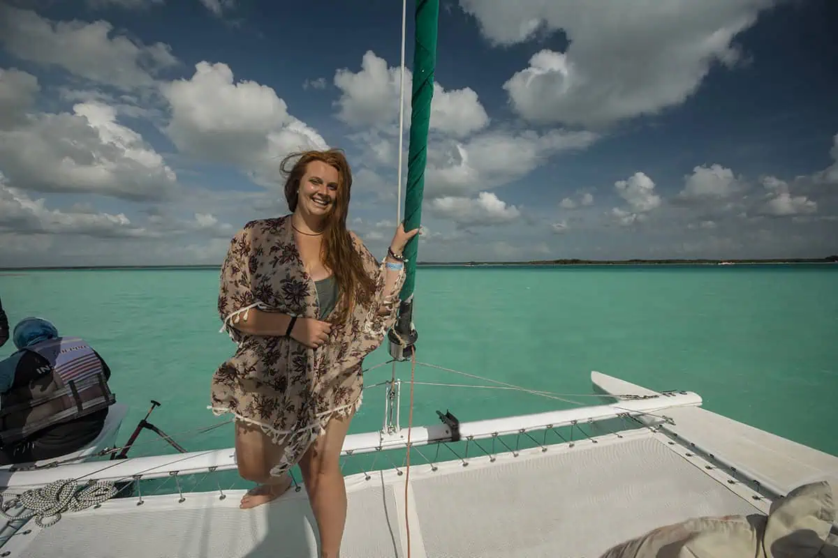 tasha amy board a sailboat on the bright turquoise waters of Bacalar. She is leaning on the netting of the catamaran, dressed in a floral dress, with her hair blowing in the wind. The sky above is partly cloudy, emphasizing the vivid color of the water.