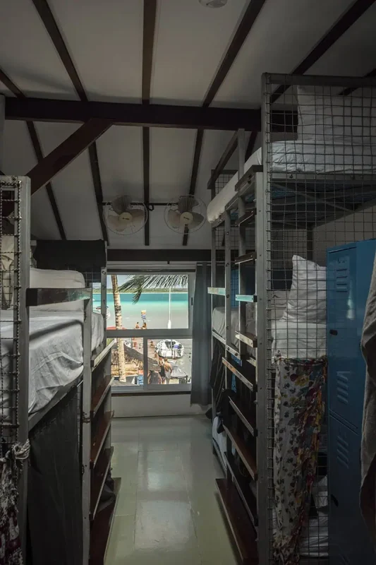Inside a yak lakehouse hostel in Bacalar, featuring a row of bunk beds with privacy curtains. The hostel's interior includes high ceilings with fans, and a large window that offers a view of a sunny beach scene outside, providing a unique and inviting atmosphere for travelers.