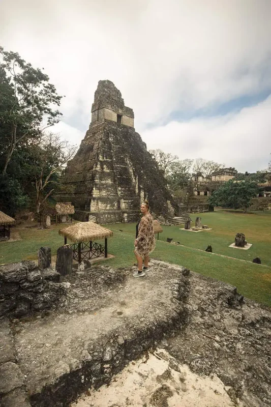 tasha amy standing in front of a massive Mayan temple at Tikal, part of a historical site near Flores. The temple rises dramatically against a backdrop of lush greenery and overcast skies