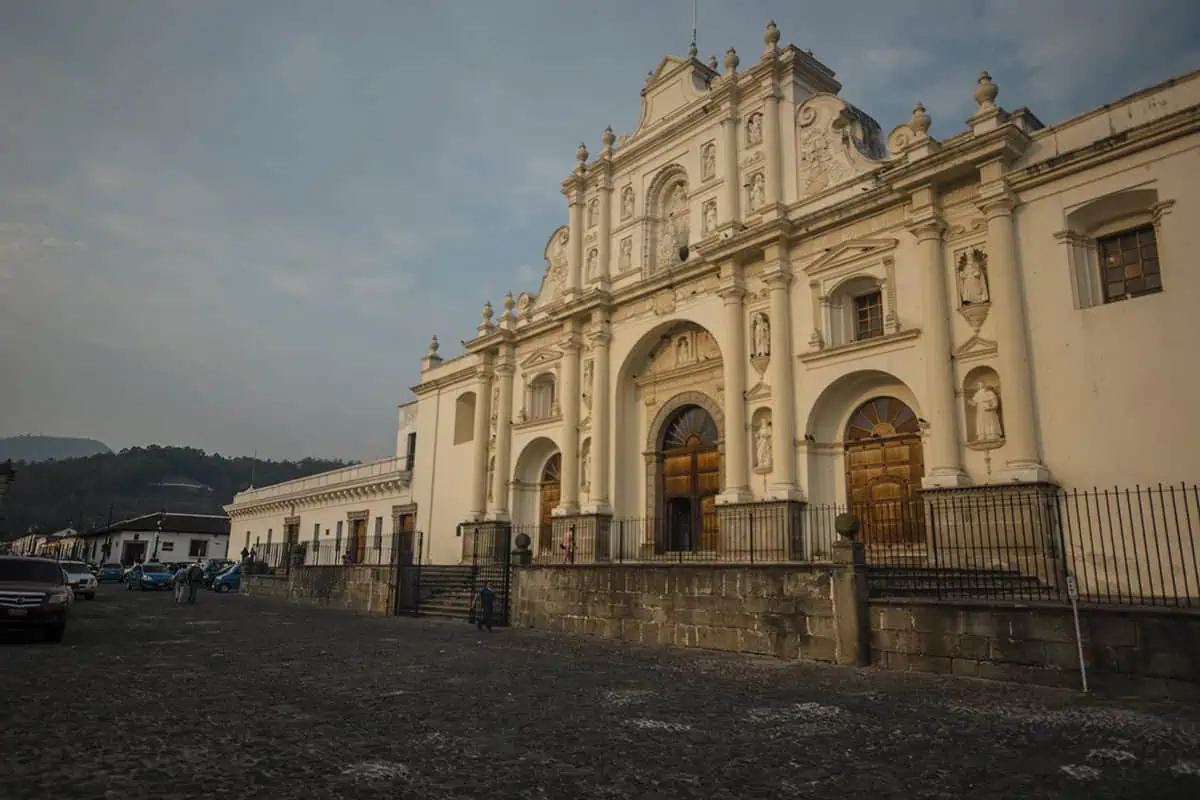 The historic Cathedral of Antigua, Guatemala, captured in the early evening with a muted sky in the background. The church's elaborate white facade features intricate sculptural details and classical architectural elements. The scene is set on a cobblestone street, enhancing the old-world charm of this colonial city.
