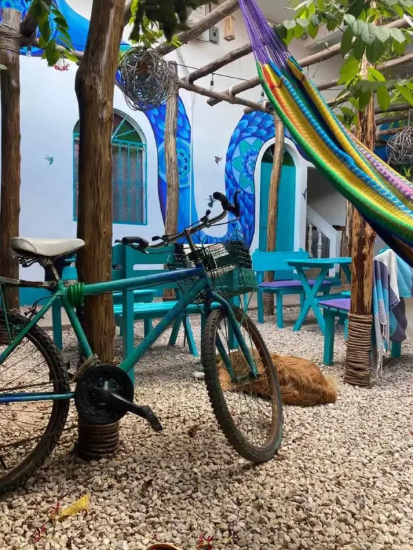 A vibrant and artistic courtyard in Flores featuring a green bicycle parked on a gravel ground, colorful hammocks, and a large mural of a blue octopus on the wall. The scene is embellished with various hanging decorations and plants, creating a lively and welcoming atmosphere.