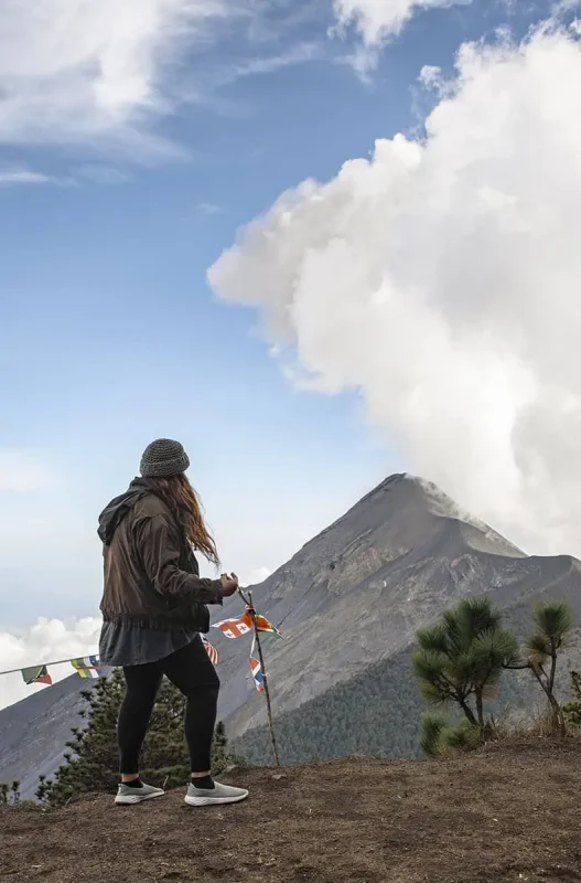 tasha amy standing on a mountain peak, observing a nearby active volcano emitting a plume of smoke in Antigua. She is dressed in outdoor gear and holding colorful prayer flags, which add a vivid touch against the rugged volcanic backdrop. This adventurous experience is a notable highlight in the Mexico, Belize, Guatemala itinerary