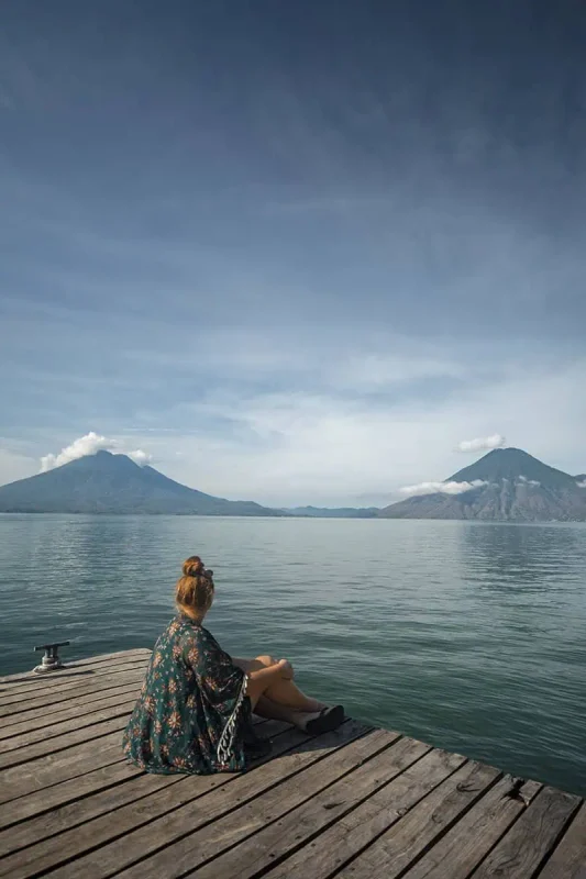 tasha amy sits on a wooden dock overlooking Lake Atitlán, surrounded by majestic volcanoes under a clear blue sky. Her contemplative pose captures a moment of peace as she gazes out over the calm waters, framed by the natural beauty of Guatemala. This picturesque setting is a serene highlight on the Mexico, Belize, Guatemala itinerary.