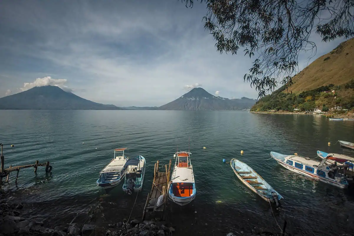 A panoramic view of Lake Atitlán, captured from a lakeside spot where boats are tied up to a rustic wooden dock. The lake is flanked by towering volcanoes and hills, with trees overhanging the water's edge, presenting a perfect blend of natural elements and serene landscapes.
