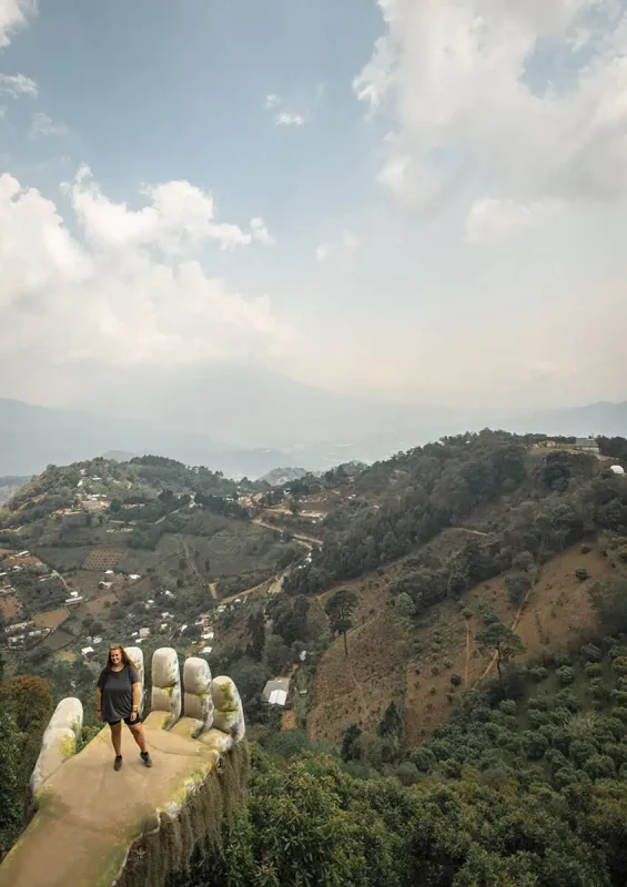 tasha amy stands on a uniquely designed viewpoint that resembles a hand, located on the edge of a hill in Antigua. Below, a panoramic view of a lush valley dotted with small buildings and patches of agriculture extends into the distance, framed by a sky filled with light clouds.