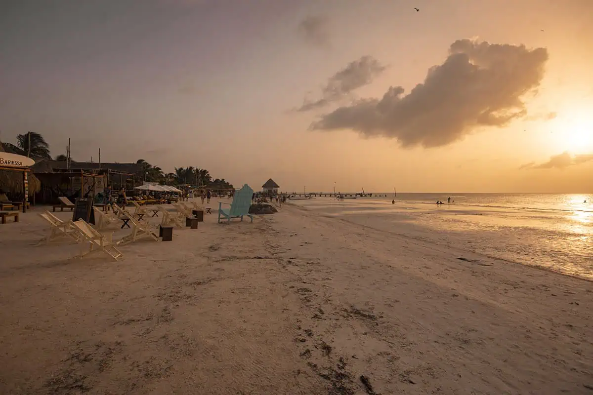A tranquil sunset view at Isla Holbox, featuring a softly lit sandy beach with scattered wooden chairs and tables. Several beachside bars and a wooden pier stretch into the calm sea, under a sky painted with hues of orange and pink. A single cloud floats above the horizon in an otherwise clear sky