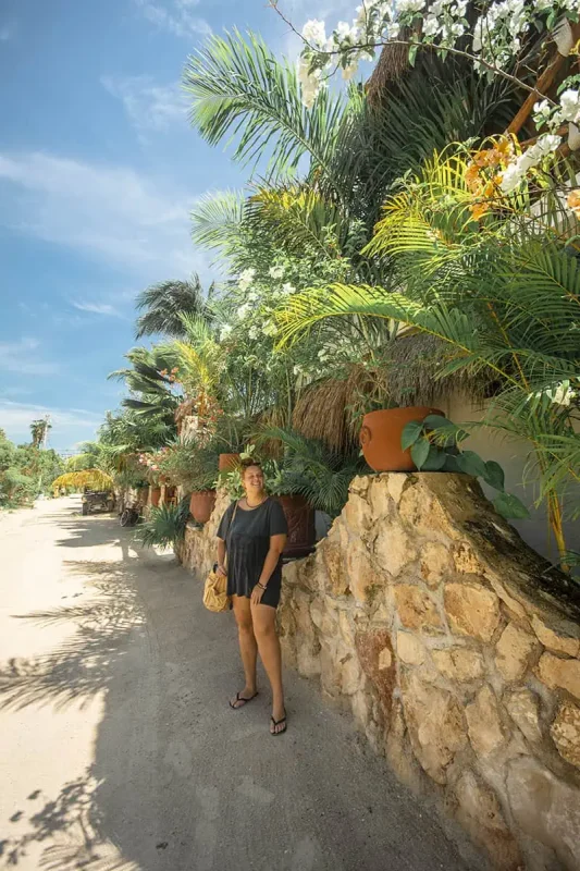 tasha amy in casual summer attire stands on a sandy pathway surrounded by lush tropical greenery and flowering plants on Isla Holbox. The path is flanked by stone walls and vibrant, densely packed tropical plants, providing a rich, natural environment.