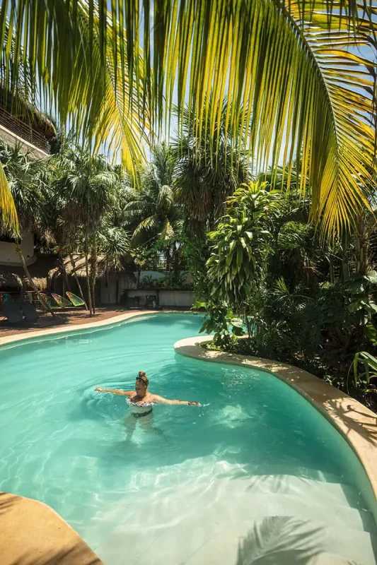 In this part of her Mexico, Belize, Guatemala itinerary, tasha amy enjoys swimming in a winding, river-like pool surrounded by dense tropical foliage on Isla Holbox. The pool's turquoise waters contrast beautifully with the vibrant green plants and the bright, sunlit setting.
