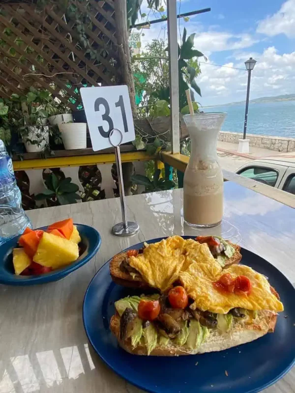 A meal set up on a beachfront restaurant table in Flores, including a hearty open sandwich topped with avocado, scrambled eggs, and tomatoes on crusty bread. A glass of iced coffee and a bowl of fresh fruit salad complement the meal, set against the backdrop of a serene ocean view, making it part of a memorable Mexico, Belize, Guatemala itinerary.