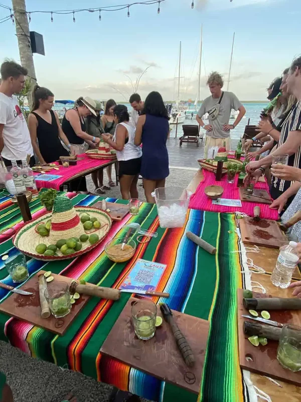 vibrant mojito-making class by the sea in Bacalar, Mexico. A group of tourists, of various ages and backgrounds, is gathered around a colorful table covered with a traditional Mexican "sarape" tablecloth. They're actively engaged in learning how to make mojitos, using ingredients like fresh limes and mint, displayed on rustic wooden chopping boards. The scene is lively and festive, set against a backdrop of sailboats moored in the turquoise waters of Bacalar Lagoon, enhancing the tropical and social atmosphere of the experience.